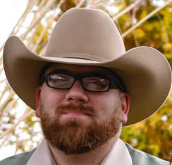Adult male with a short read beard, black rectangular glasses and a tan cowbow hat staring off into the distance with a slight smile